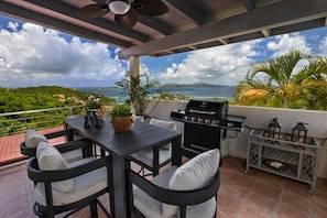 A patio with a table and chairs, a grill, and a view of the ocean and hills.