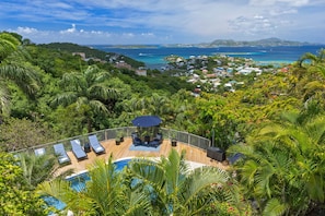 A pool with lounge chairs and a table, surrounded by lush greenery and overlooking a coastal town.