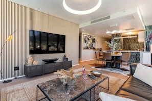 Un salon moderne doté d’un téléviseur à écran plat, d’un mur décoratif en bois, d’un coin repas avec des chaises et d’une cuisine en arrière-plan.