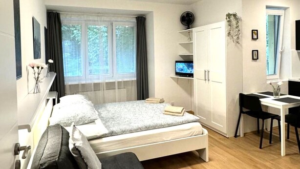 Un dormitorio moderno con cama, escritorio y silla. Hay una ventana con cortinas, un ventilador y un televisor en una estantería.