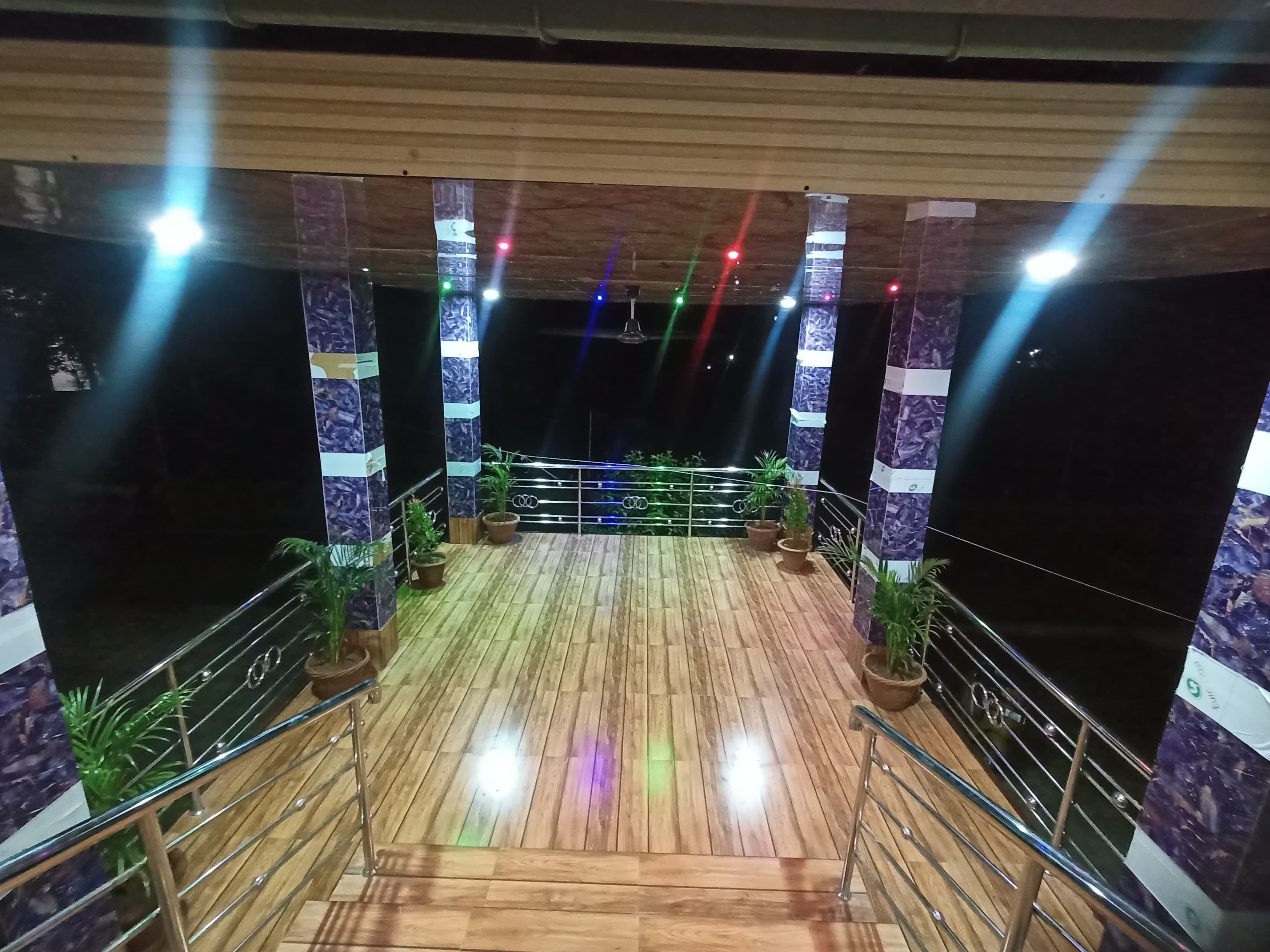 A wooden deck with railings, potted plants, and a stage with colorful lighting.
