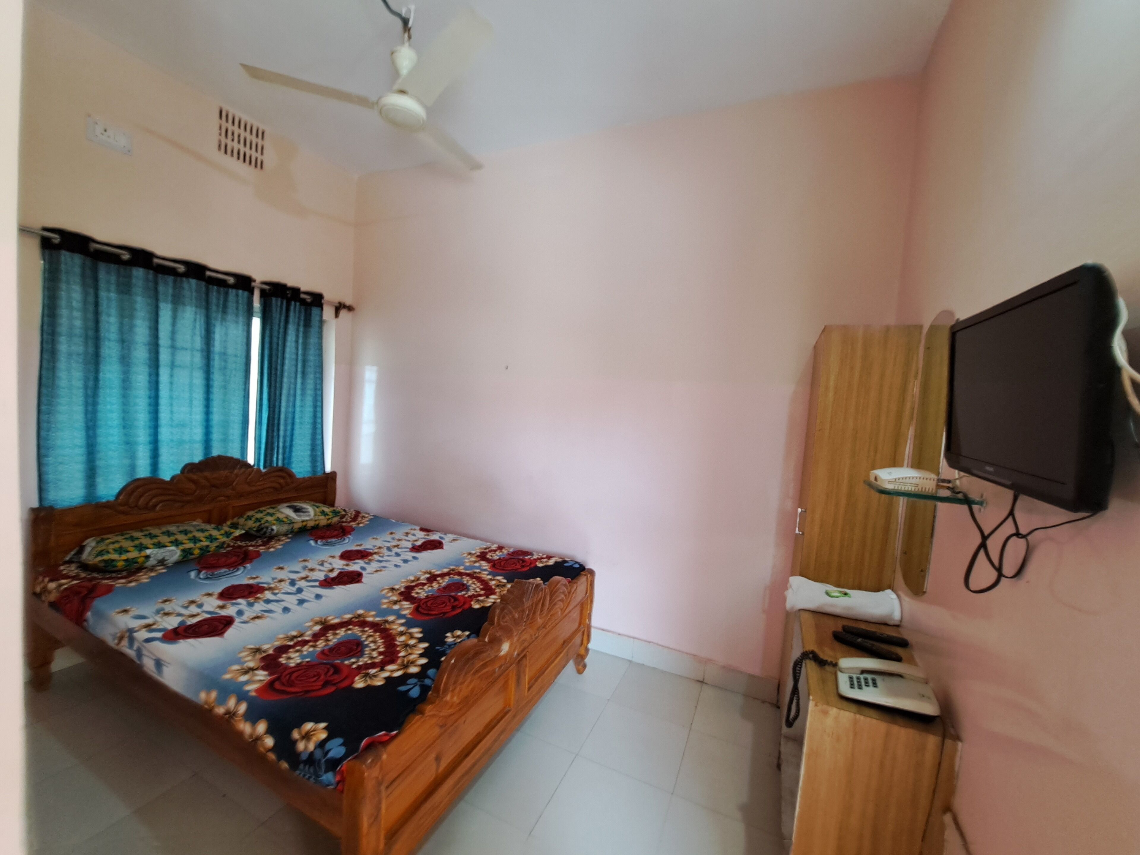 A bedroom with a bed, a wooden wardrobe, a television mounted on the wall, and a ceiling fan.