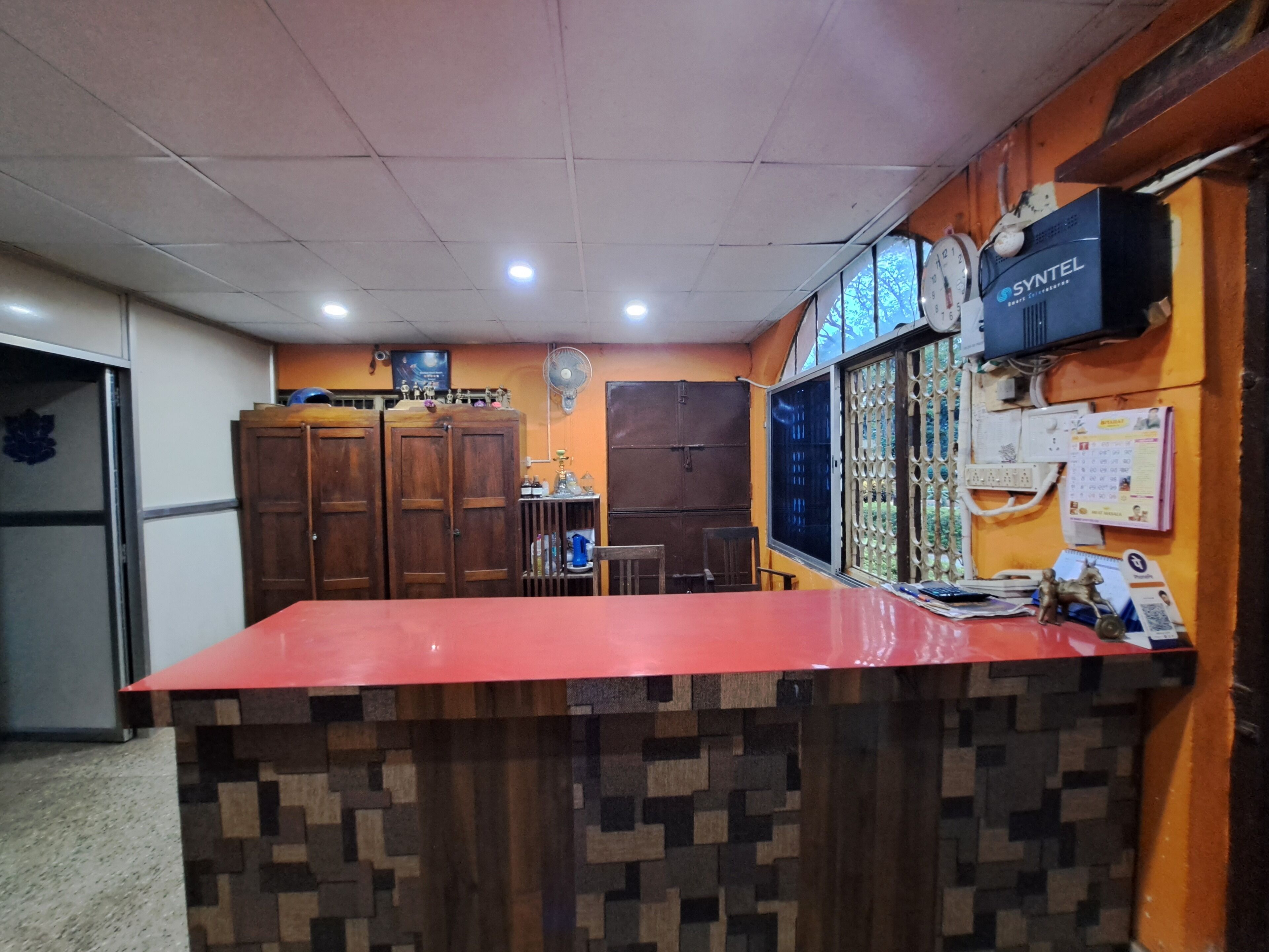 Un área de recepción con un mostrador rojo, armarios de madera, un ventilador y un reloj en la pared.