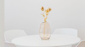 A white dining table with a clear glass vase containing dried flowers. Three framed abstract artworks are hung on the wall above the table.