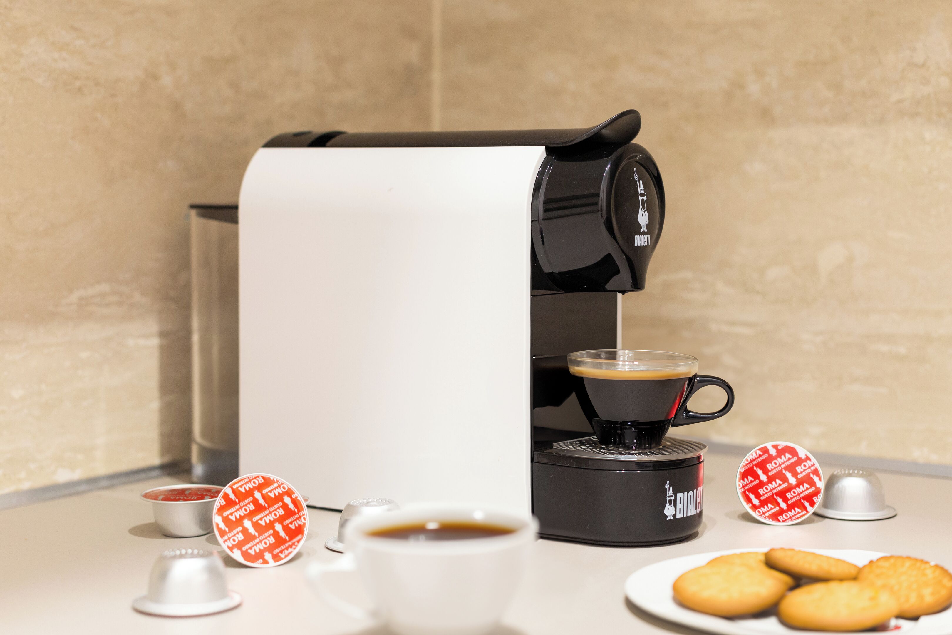 A coffee machine dispensing coffee into a cup, with a plate of cookies and a small bowl of condiments on a table.