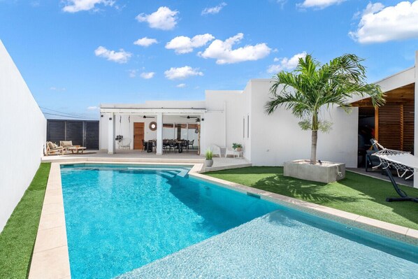 Un área de piscina moderna al aire libre, con una piscina azul cristalina, un patio con asientos y una zona de comedor cubierta.