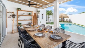 A dining area with a pool view, featuring a wooden table, black chairs, and a thatched roof.