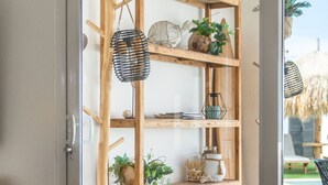 A dining area with a wooden table set for a meal, a wooden shelf with decorative items, and a view of the outdoors.
