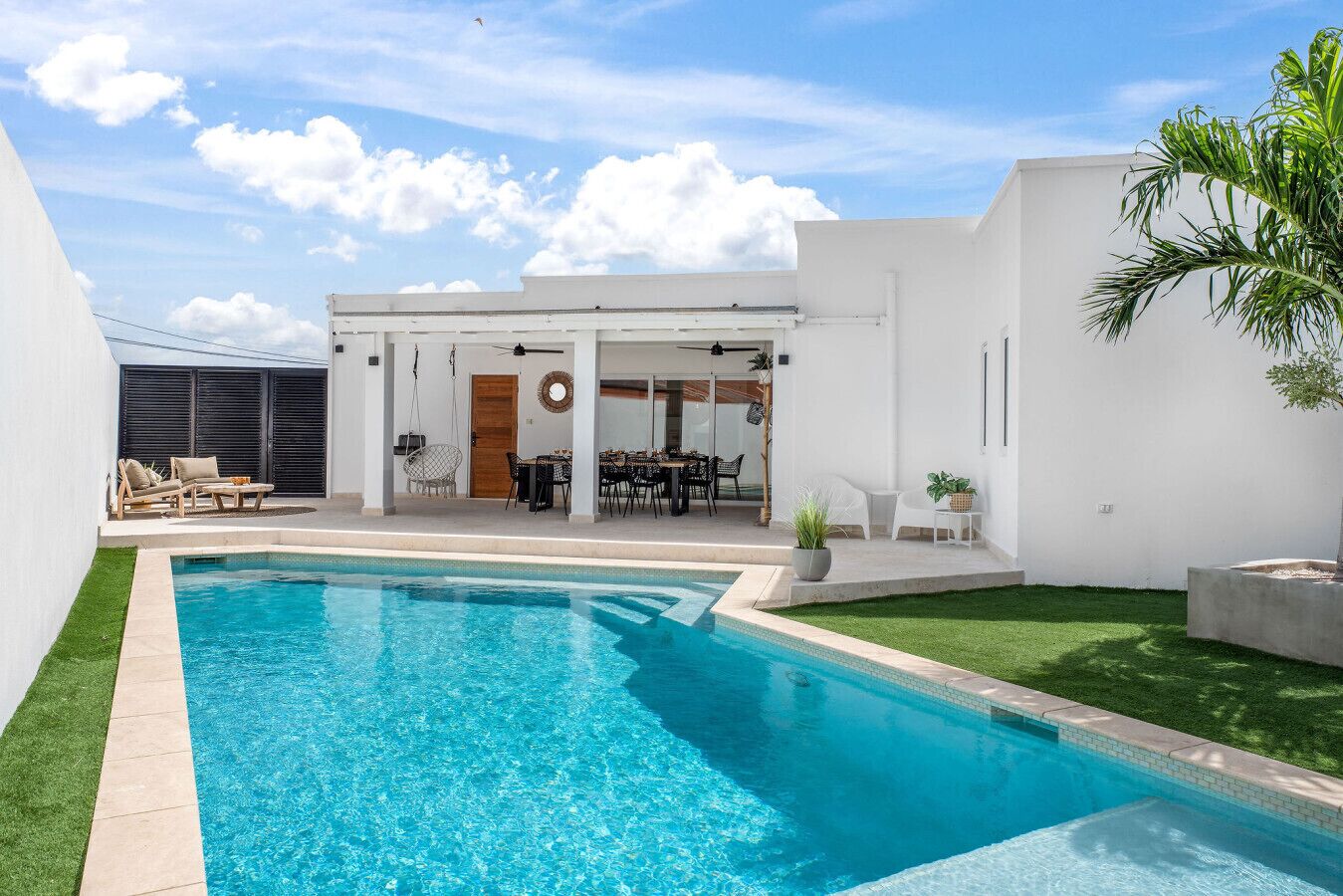 Un’area piscina moderna con piscina blu cristallina, un patio con mobili da esterno e un edificio bianco dotato di portico coperto.
