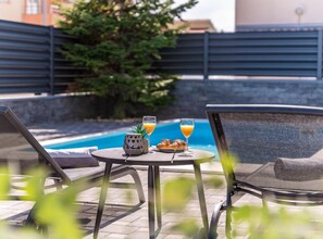 Un espace aménagé au bord de la piscine avec deux chaises longues, une table sur laquelle se trouvent un verre de jus d’orange, un verre d’eau et un croissant.