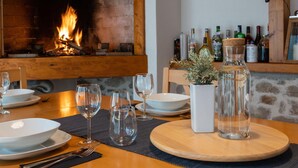 Mesa de comedor dispuesta con platos, vasos y cubiertos, una planta en maceta y una botella de agua. Se ve una chimenea con fuego encendido al fondo.