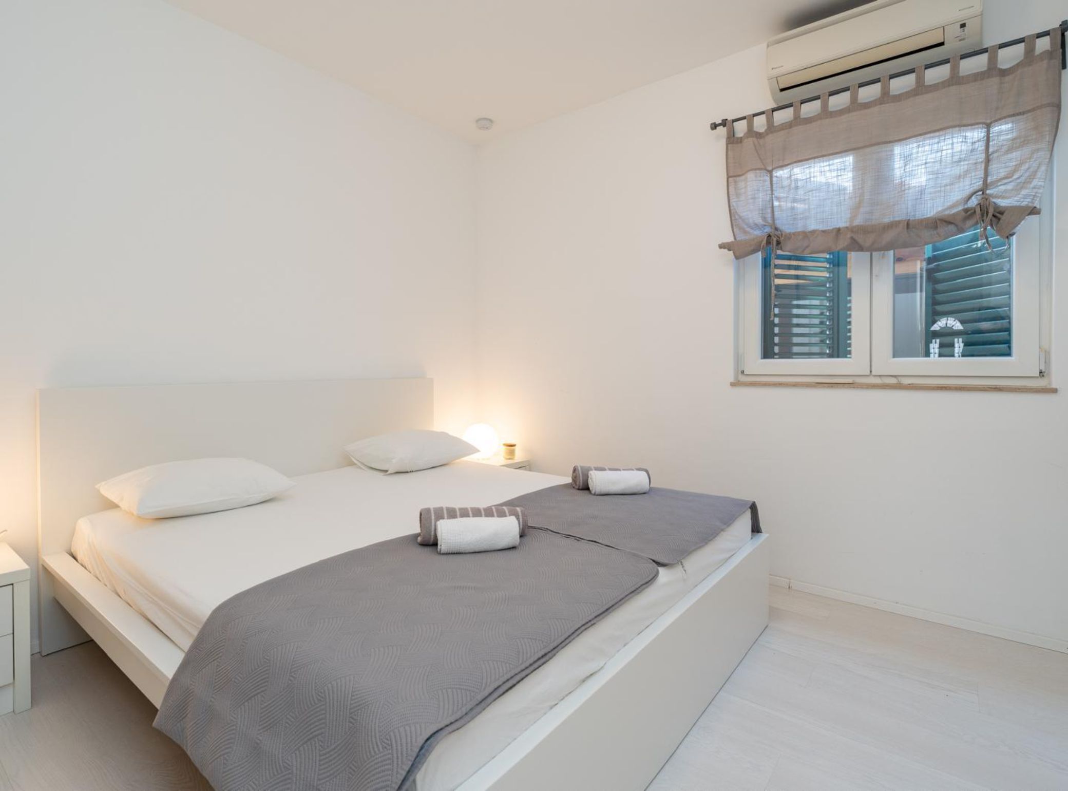 Un dormitorio con cama, mesitas de noche, ventana con persianas y aire acondicionado.