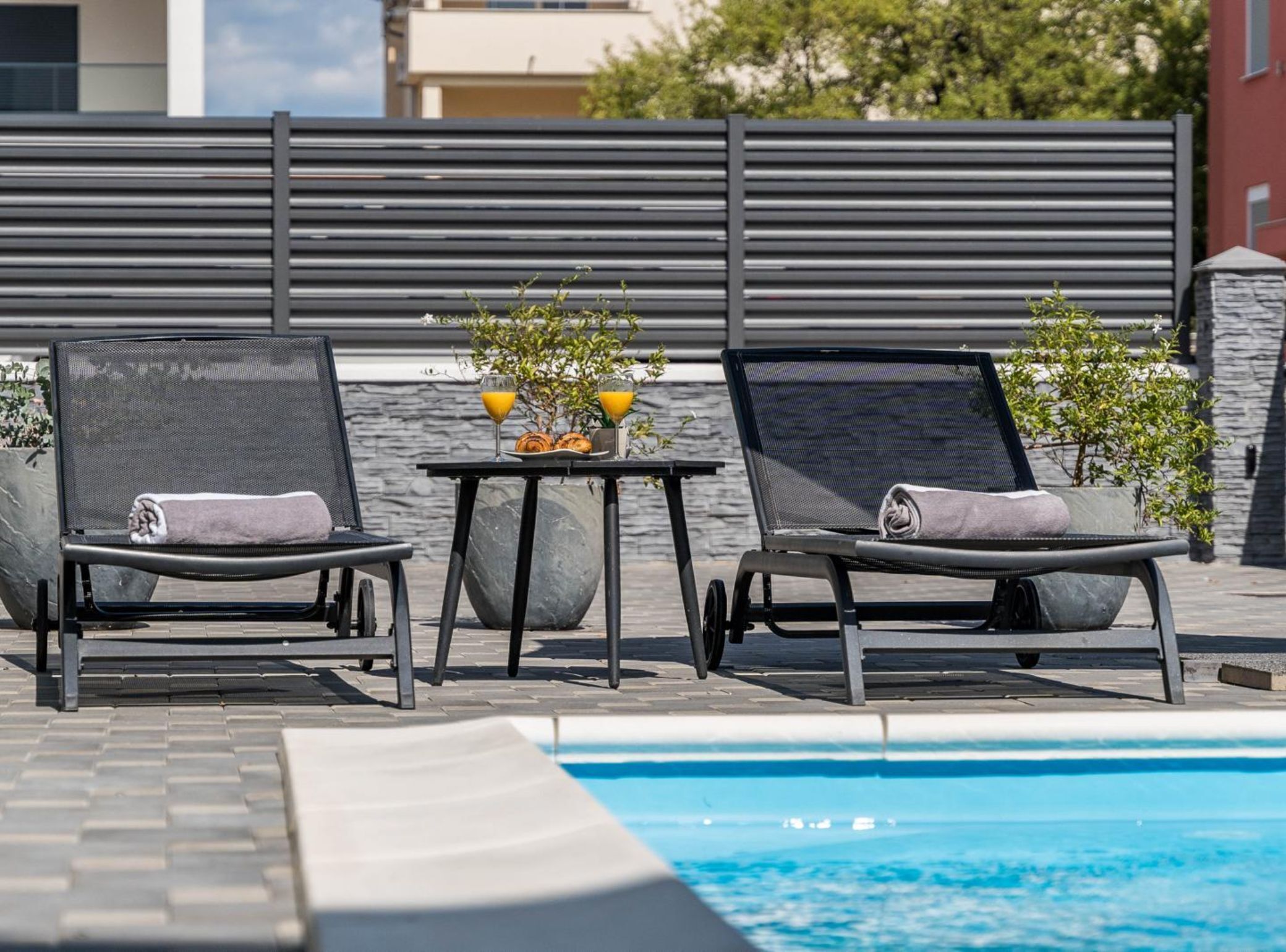 Dos sillones de relax junto a una piscina, con una mesa y bebidas, frente a una fachada de edificio moderno.