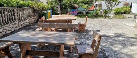 An outdoor seating area with wooden tables and benches, surrounded by a stone wall and greenery.