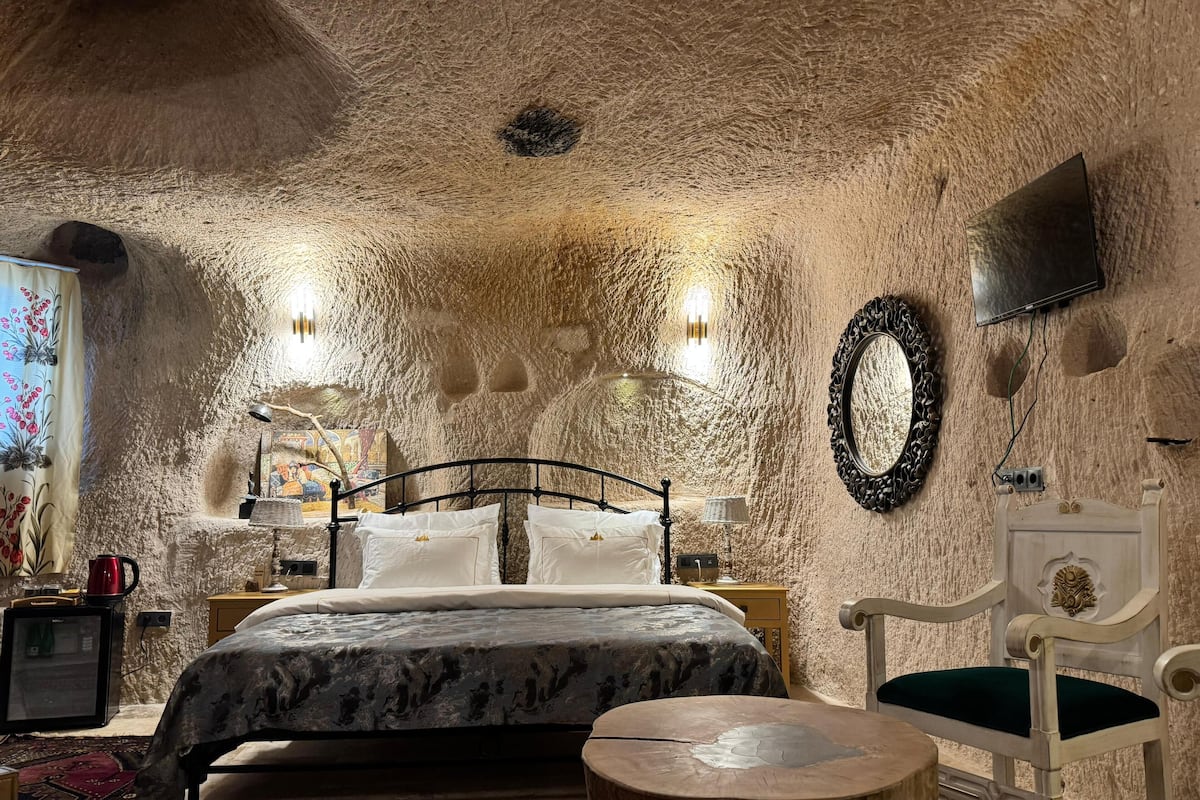 Un dormitorio con pared de piedra, una cama con ropa de cama blanca, una mesita de madera, una silla y un televisor montado en la pared.