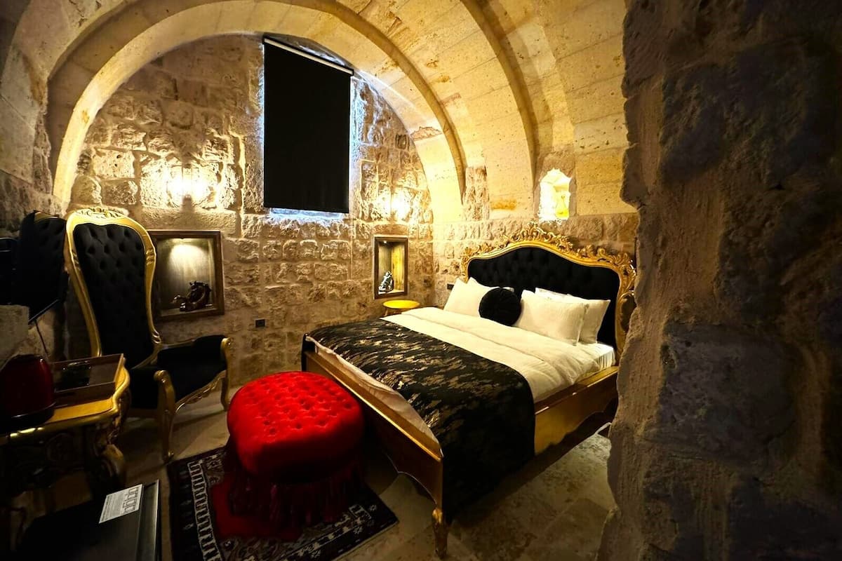 Un dormitorio con pared de piedra, ventana arqueada, cama con cabecero negro, un reposapiés rojo y una pequeña mesa con un teclado.