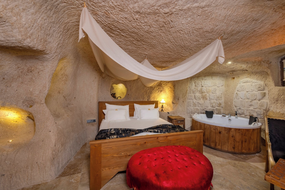 A bedroom with a wooden bed, a red ottoman, a stone wall, and a stone bathtub.