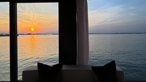 A room with a large window overlooking a sunset over water, a sofa with cushions, and a curtain.