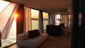 A modern interior with a sofa and a dining area, large windows with curtains, and a view of the water.