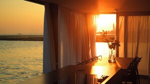 A room with a view of the sunset over water, a table with a vase of flowers, and a chair.