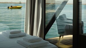 A bedroom with a large window overlooking a body of water, a bed with white linens, and a chair by the window.