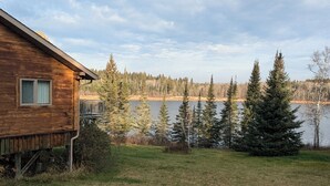 Una cabaña de madera con terraza que da a un lago, rodeada de árboles y zonas verdes.