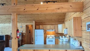 Interior de una cabaña de madera con una zona de cocina que incluye un refrigerador, estufa y taburetes de bar.