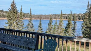 Una terraza de madera con una silla Adirondack verde y un banco, con vistas a un lago y árboles al fondo.
