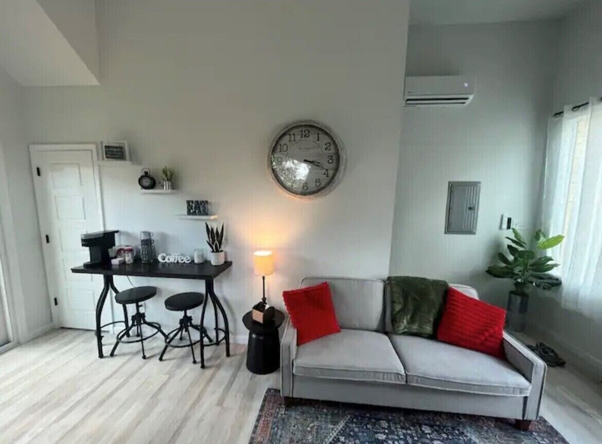 Ein Wohnzimmer mit einer Couch, einem Couchtisch, einer Uhr und einer kleinen Küchenzeile mit Hockern.