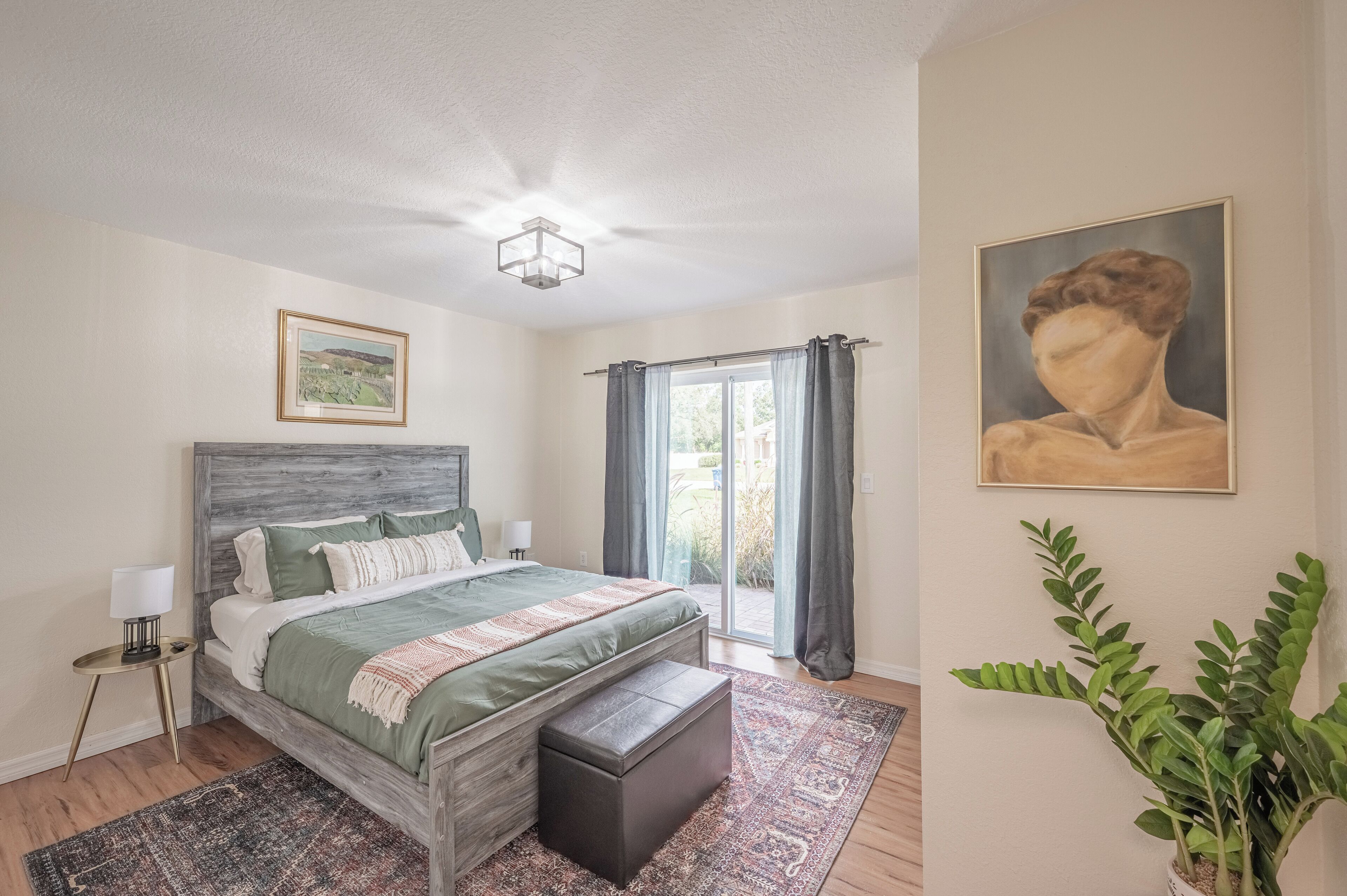 A bedroom with a bed, a nightstand, a painting, and a potted plant.
