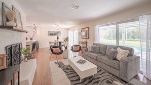 A spacious living room with a grey sofa, a white coffee table, and a zebra-patterned rug. There is a dining area with a table and chairs, and a large window offering a view of the outdoors. - Weeki Wachee Pod


 (Spring Hill)
