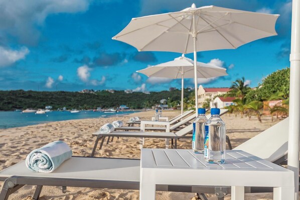 Um espaço de relaxamento à beira-mar, com guarda-sóis brancos, espreguiçadeiras e uma mesa com água engarrafada.