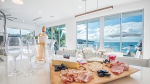 A well-lit room with a large window overlooking the sea, a wooden board with various cheeses and meats, and several wine glasses. - Luxury Beachfront Enclave Unit 1 (Sandy Ground)