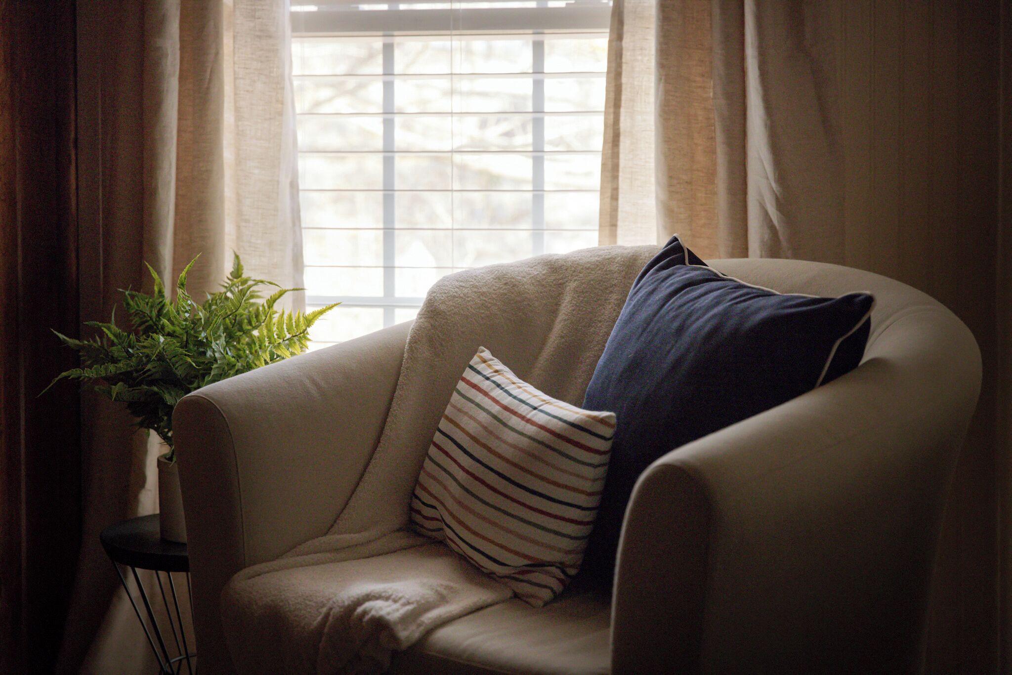 A beige armchair with striped and solid blue pillows, positioned near a window with blinds.