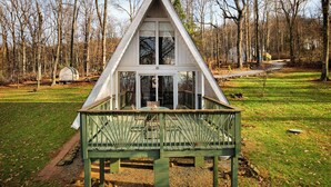 A triangular wooden structure with a glass front and a green deck, surrounded by a grassy area and trees.