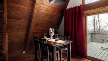 A dining area with a wooden table set for four, a chandelier, and a view of the deck through a large window with red curtains.