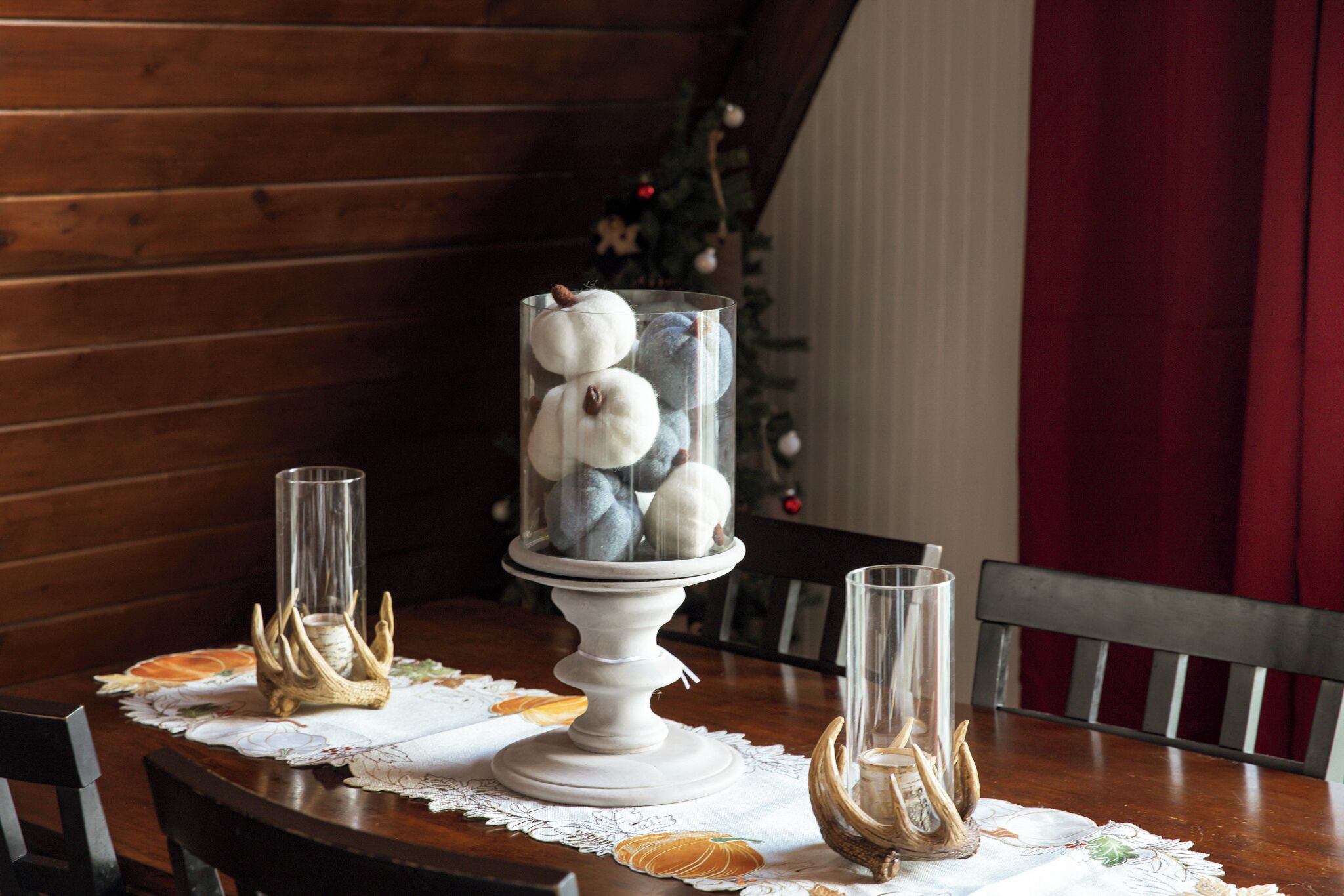 A dining table with a white lace tablecloth, two glass candle holders, and a decorative glass vase with pumpkins.