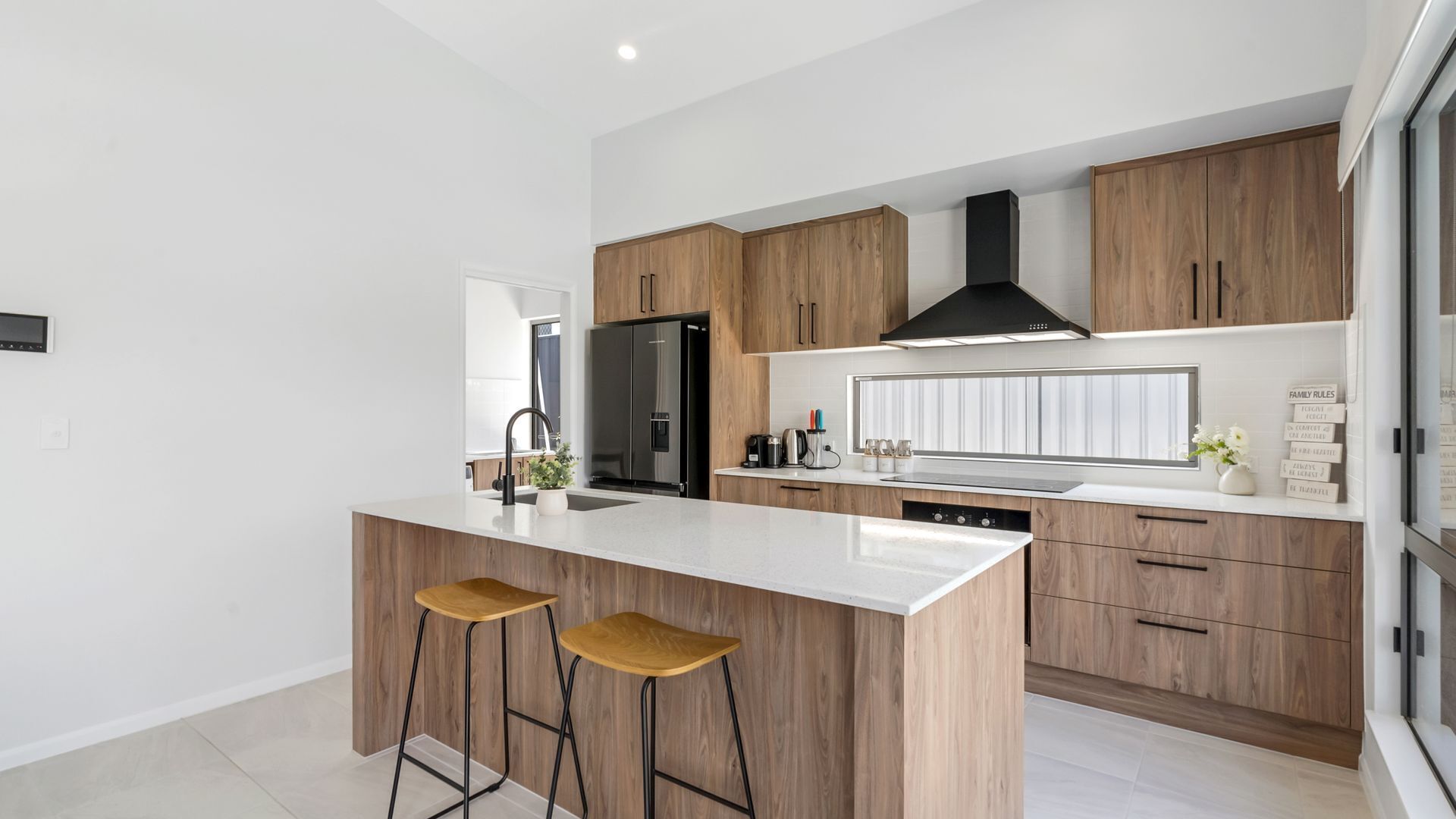A modern kitchen with a central island, wooden cabinets, a black range hood, and a stainless steel refrigerator.