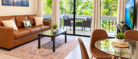A living room with a sofa, coffee table, and chairs, a TV, and a balcony with outdoor furniture.