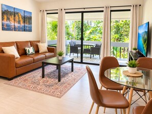 A living room with a sofa, coffee table, and chairs, a TV, and a balcony with outdoor furniture.