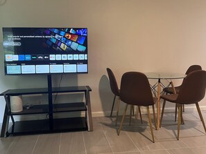A flat-screen TV on a stand with a home dashboard interface, a glass-top dining table, and two brown chairs. - Newly Renovated Cozy 2Br 1Ba Apt in Waiakea Villas (Hilo)