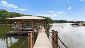 A wooden dock leading to a covered seating area with a boat and a small pavilion. - Whitetail Cove by Avantstay Lakefront w/ Pool + Private Dock Sleeps 20 (Troutman)