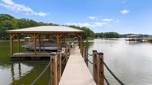 A wooden dock leading to a covered seating area with a boat and a small pavilion. - Whitetail Cove by Avantstay Lakefront w/ Pool + Private Dock Sleeps 20 (Troutman)