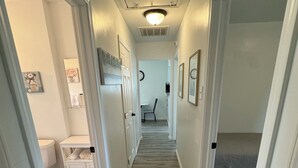 Un pasillo estrecho con suelo de madera, paredes blancas y puertas que dan a diferentes habitaciones. Se observan cuadros enmarcados en las paredes, un reloj y una pequeña mesa con una silla al fondo.