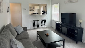Una sala de estar con un sofá gris, una mesa de centro negra, un televisor sobre un soporte negro, y un área de cocina con una encimera y gabinetes blancos.
