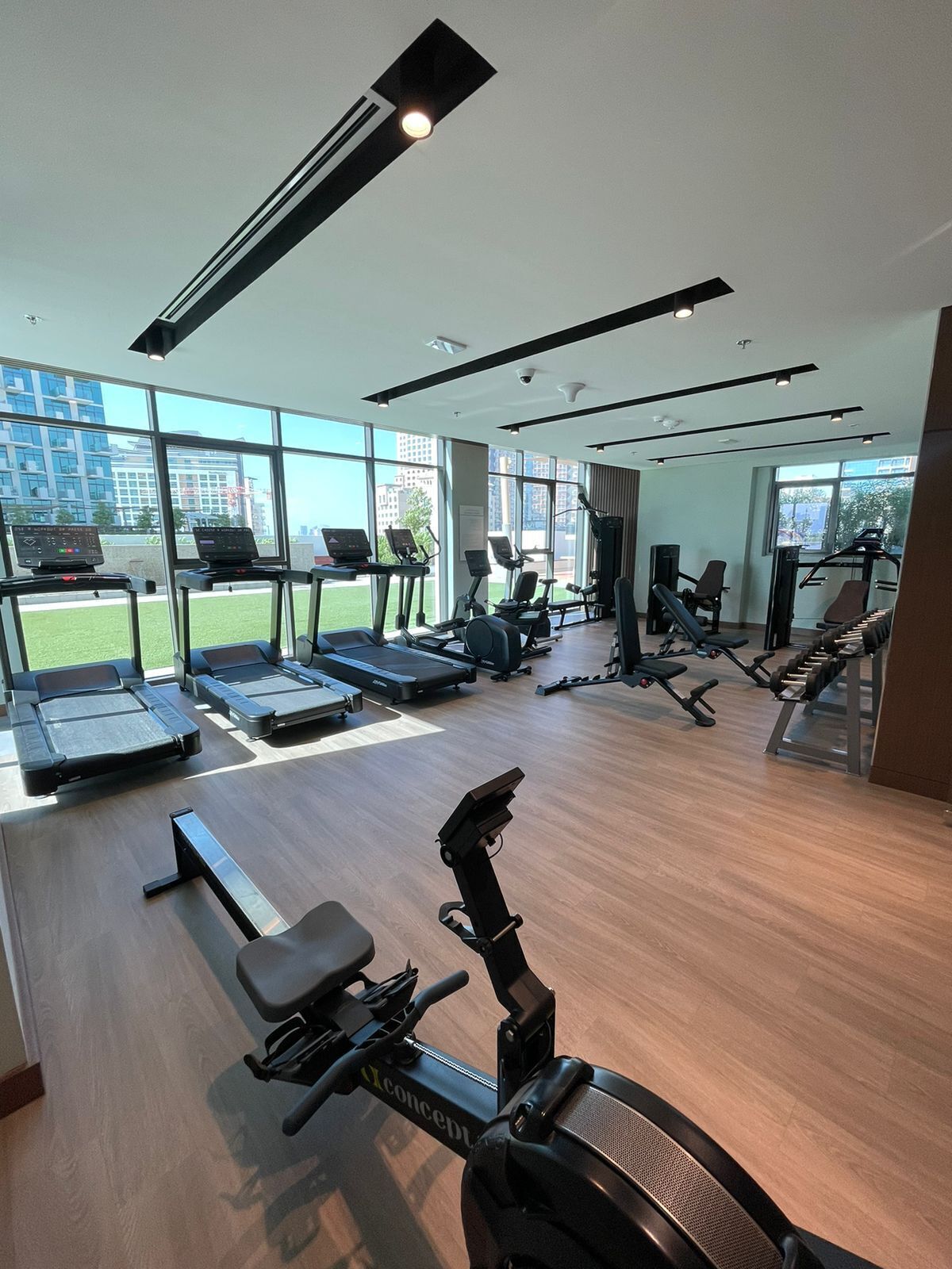 A modern gym with treadmills, elliptical machines, and weight benches. Large windows offer views of the cityscape.