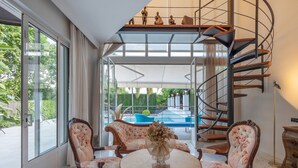 Una habitación amplia con un gran ventanal, una mesa redonda con sillas, un jarrón decorativo, una escalera de caracol y vistas a una piscina.