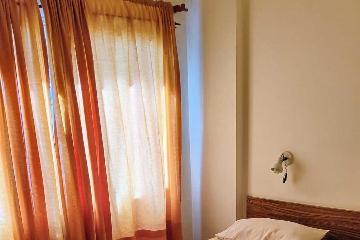 A single bed with a beige bedspread and a pillow, a wooden headboard, a bedside table, and a wall-mounted light fixture.