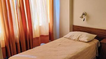 Una cama individual con edredón color beige y una almohada, cabecera de madera, mesita de noche y aplique de pared.