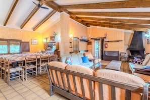 A cozy living room with a high ceiling, wooden beams, a fireplace, and a dining area with a large table and chairs. - Rural House 'Can Guidic' with Mountain Views, Private Pool and Wi-Fi (La Vall D' en Bas)